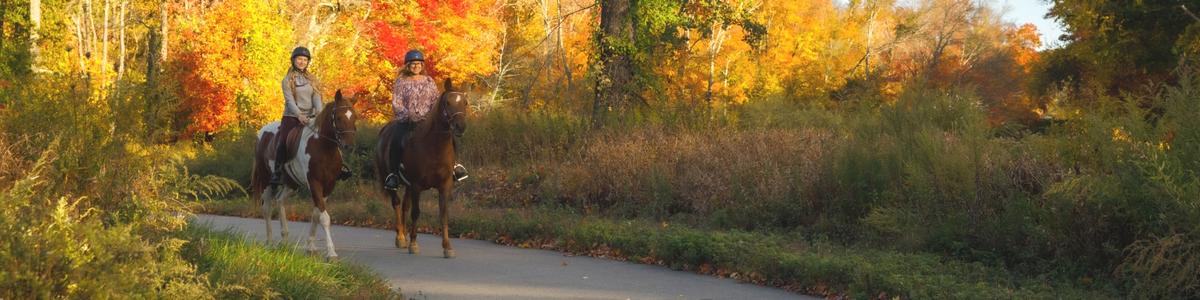 Two women ride their horses on a paved path surrounded by fall colors.