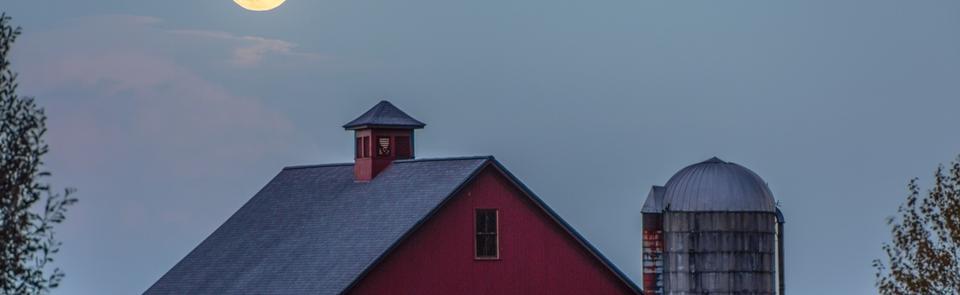 A red barn at night under the moon to the left of a silver silo.