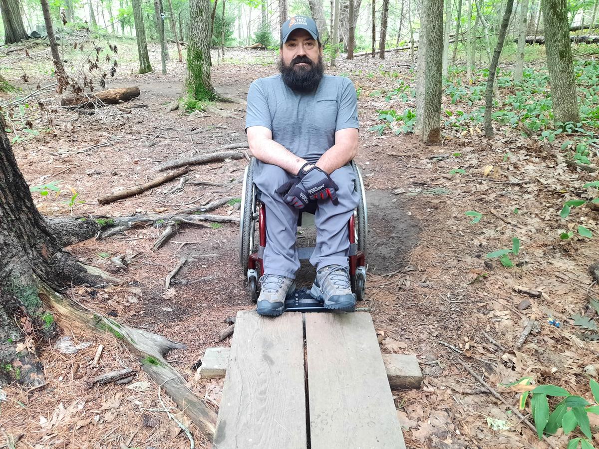 A man in a wheelchair sits next to a bog bridge that is two narrow for his chair.