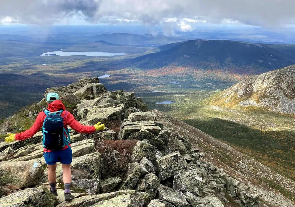 A woman hikes down a rocky ridge with views of mountains around her