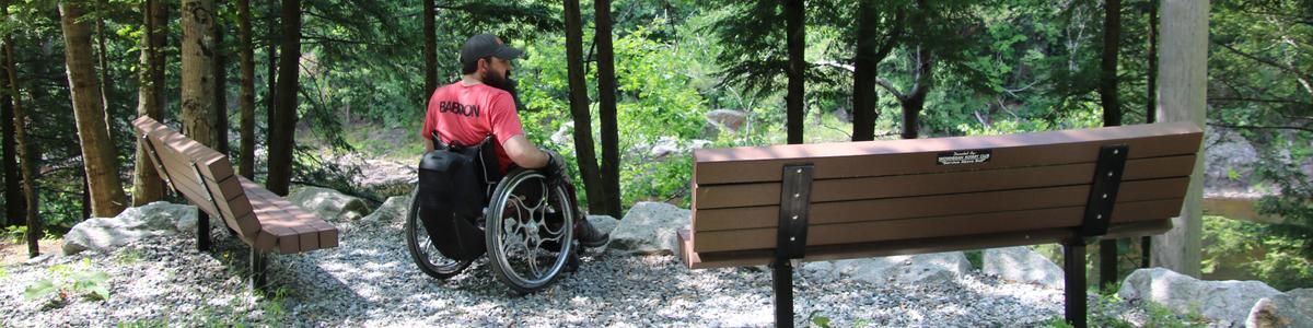 A man in a wheelchair wearing a red shirt and gray cap sits looking out over the Kennebec River