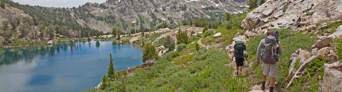 Two backpackers hiking around a mountain lake with jagged mountain peaks visible