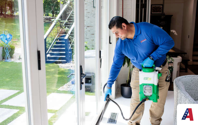 A pest control technician in a blue uniform points into an open crawl space with a flashlight while explaining a pest issue to a female homeowner outside a brick house.