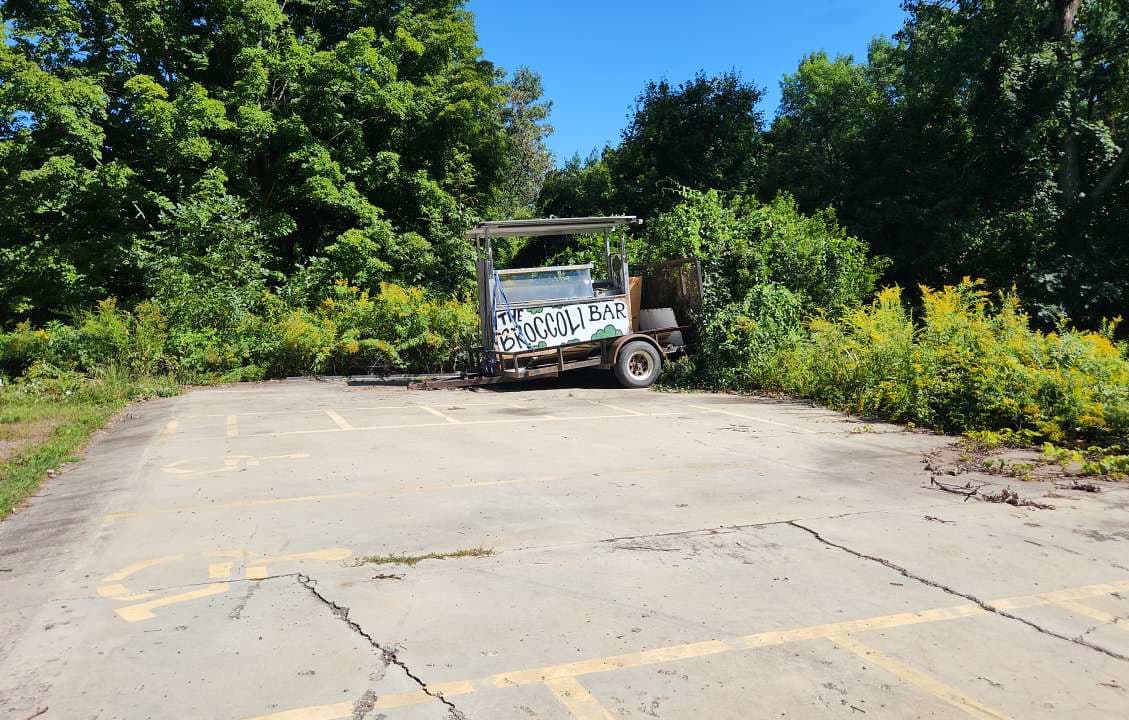 Paved, cracked parking area with handicapped spaces delinated and falling apart "Broccili Bar" cart