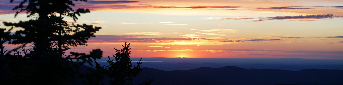 The sun peeks over the silhouettes of rolling hills in a valley, turning the sky and clouds gold, pink, and purple.